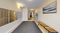 This 28 square meter space serves as the lobby of an apartment building, efficiently designed to maximize functionality in a compact area. The entrance is highlighted by a large glass door, offering natural light and picturesque views of snow-covered steps and landscaping. Inside, the lobby boasts a clean, modern aesthetic with cream-colored walls and a combination of tile and carpeted flooring.

Security is a priority, evident from the numerous mailboxes lining one wall and a prominently displayed security camera notice. The lobby houses an elevator for convenient access to upper floors, complete with safety signage. Lighting is well-considered, with a mix of ceiling fixtures and wall sconces creating a bright, welcoming atmosphere.

The space is thoughtfully furnished with a wooden bench for seating and a decorative plant, adding warmth to the functional design. A large abstract artwork adorns one wall, introducing a splash of color to the neutral palette. The layout efficiently accommodates both utility and comfort, serving as a transitional space between the outside world and private residences.