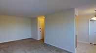 This single-floor space presents a clean, functional layout with a straightforward design aesthetic. The entry area is defined by a solid white door with a classic knob, set within a simple white-trimmed frame. Mounted on the adjacent wall is a built-in intercom panel with multiple controls, adding a practical communication feature to the entryway. The walls throughout are finished in a soft, neutral tone that contributes to a calm and understated ambiance.

The main living area features wall-to-wall carpeting in a muted, earthy tone, providing a comfortable and quiet underfoot experience. White baseboards line the perimeter, offering a clean contrast against the neutral walls. The open layout of the room allows for flexible furniture arrangement, and a doorway connects the living space back to the entry corridor, creating a cohesive flow between areas. Electrical outlets are conveniently positioned along the walls. Overhead lighting in the entry corridor casts a warm glow into the main room. Overall, the space maintains a simple, move-in-ready character with minimal ornamentation.