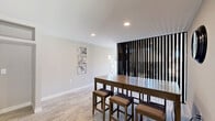 Located steps from the Atlantic Ocean at 1500 S Ocean Blvd in Myrtle Beach, SC, this thoughtfully designed single-floor space functions as a combined fitness room and social lounge. The main area features light wood-look flooring throughout, recessed lighting, and crisp white walls that create a clean, modern aesthetic. A striking dark vertical wood slat accent wall serves as a dramatic focal point, dividing the space with bold visual contrast. The fitness zone is equipped with a stationary spin bike and a rowing machine, bathed in abundant natural light through a floor-to-ceiling glass corner window that frames views of the surrounding palm-lined outdoor area. A gallery-style arrangement of mirrored panels on the adjacent wall enhances the sense of space in the workout area. The lounge area features a dark wood high-top bar table with cushioned stools and is complemented by abstract artwork displayed on the walls. A hallway connects to the bathroom, which is finished with a distinctive pebble-stone mosaic floor, a floating dark vanity, a round rope-hung illuminated mirror, and a decorative surfboard wall accent — a nod to the coastal location. The overall ambiance is contemporary, coastal, and functional.