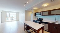 This single-floor residence is thoughtfully designed with a contemporary aesthetic and a practical open-concept layout. The kitchen serves as a central highlight, outfitted with rich dark-wood cabinetry featuring frosted glass inserts, a light-colored tile backsplash, and a full suite of stainless steel appliances including an electric range, built-in microwave, dishwasher, and refrigerator. A freestanding island with a white countertop and built-in wine storage adds both workspace and visual interest. Track and flush-mount lighting illuminate the kitchen and adjoining living area, which is finished with large-format light tile flooring and flows seamlessly throughout the main living space. A stacked washer and dryer unit is conveniently located within the main living area. The bedrooms feature neutral carpet and receive generous natural light through large windows that frame urban views of the surrounding cityscape. Two bathrooms serve the unit: one with a glass-enclosed walk-in shower and dark wood vanity, and a second with a full soaking tub, tiled surround, and matching cabinetry. The overall palette of cream walls, light flooring, and warm wood tones creates a clean, modern, and inviting atmosphere throughout.