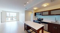 This single-floor residence is thoughtfully designed with a contemporary aesthetic and a practical open-concept layout. The kitchen serves as a central highlight, outfitted with rich dark-wood cabinetry featuring frosted glass inserts, a light-colored tile backsplash, and a full suite of stainless steel appliances including an electric range, built-in microwave, dishwasher, and refrigerator. A freestanding island with a white countertop and built-in wine storage adds both workspace and visual interest. Track and flush-mount lighting illuminate the kitchen and adjoining living area, which is finished with large-format light tile flooring and flows seamlessly throughout the main living space. A stacked washer and dryer unit is conveniently located within the main living area. The bedrooms feature neutral carpet and receive generous natural light through large windows that frame urban views of the surrounding cityscape. Two bathrooms serve the unit: one with a glass-enclosed walk-in shower and dark wood vanity, and a second with a full soaking tub, tiled surround, and matching cabinetry. The overall palette of cream walls, light flooring, and warm wood tones creates a clean, modern, and inviting atmosphere throughout.