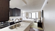 This single-floor studio suite offers a thoughtfully designed open layout that efficiently combines living, sleeping, and cooking areas into one cohesive space. The interior features a neutral warm palette of beige and white tones complemented by dark espresso cabinetry and hardwood-style flooring, creating a modern yet inviting atmosphere.

The fully equipped kitchen is a standout feature, outfitted with dark upper and lower cabinetry, stainless steel appliances including a full-size refrigerator, electric range, and microwave, plus a marble-style countertop with an undermount sink. A built-in wine rack adds a refined touch to the kitchen area.

The sleeping area accommodates a large bed dressed in crisp white linens, positioned to face a wall-mounted flat-screen television atop a dark media console. Framed artwork above the headboard adds a decorative accent, while crown molding details the textured ceiling throughout.

A windowed seating nook provides a bright corner retreat with a loveseat and accent chair, offering views of the surrounding cityscape through large horizontal-blind-fitted windows. Natural light filters generously into the space.

The bathroom is clean and functional, featuring a dark vanity with a marble-style countertop and wood-look flooring, maintaining the suite's consistent design aesthetic.