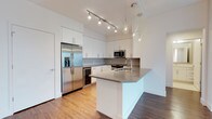 This single-floor 2-bedroom, 2-bathroom residence offers a thoughtfully designed layout with modern finishes and an open, airy feel. The kitchen serves as a standout centerpiece, showcasing a clean contemporary aesthetic with bright white cabinetry that extends to the ceiling for generous storage. A large kitchen island with a gray quartz countertop provides ample prep space and doubles as a casual dining or entertaining area. Stainless steel appliances — including a French door refrigerator with an in-door ice and water dispenser, a built-in microwave, and a dishwasher — complete the well-appointed culinary space. Pendant lighting above the island and a multi-head track light system overhead ensure the kitchen is well-lit and inviting. Warm wood-tone flooring flows throughout, creating a cohesive and welcoming atmosphere. The open-concept design allows natural sightlines across the living areas, enhancing the sense of space. With two full bathrooms and two bedrooms, this residence offers a functional and stylish layout suited to a variety of lifestyles. The overall design balances practicality and modern elegance, making it a well-rounded and comfortable living space.