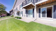 This ground-floor residence is part of a well-maintained multi-story building that showcases a classic brick exterior complemented by crisp white trim and navy blue shutters. The façade exudes a refined, traditional aesthetic, with the upper floors featuring balconies framed by black metal railings and warm wood-paneled ceilings, adding architectural depth and visual interest to the structure.

The unit benefits from expansive sliding glass doors that flood the interior with abundant natural light while providing seamless access to a private concrete patio. The patio extends directly onto a lush, meticulously maintained lawn, creating a smooth transition between indoor and outdoor living. Thoughtfully placed landscaping, including ornamental shrubs and decorative stone accents, lines the building's base, enhancing the overall curb appeal.

The building's exterior is clad in light-toned brick, lending durability and a timeless character to the property. Generous windows throughout ensure that natural light permeates the space, while the surrounding greenery contributes to a tranquil, park-like setting. The combination of quality materials, careful landscaping, and well-considered architectural details makes this a distinguished and comfortable residence suited for those who appreciate both style and practicality.