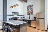 Located at 317 E Wilson St in Madison, WI, this distinctive loft-style residence showcases the character of an industrial conversion through its striking architectural details. The open-concept living and kitchen area is anchored by soaring ceilings with original exposed timber beams, raw ductwork, and large windows that flood the space with natural light. A full exposed brick wall adds texture and warmth, while light hardwood flooring runs throughout, creating a cohesive aesthetic. The kitchen features stainless steel appliances, a subway tile backsplash, dark cabinetry, and a breakfast bar peninsula. A long, well-lit hallway connects the living area to the three bedrooms, its white walls and arched mirror contrasting beautifully with the dark-framed doors and the rustic ceiling above. Each bedroom is thoughtfully appointed with light hardwood floors, clean white walls, and ample closet space with wire shelving. One bedroom includes a convenient in-unit stacked washer and dryer. The bathroom offers a clean, modern design with dark marble-style tile flooring, a walk-in glass shower, and a wood-accented vanity with a white countertop. This space masterfully blends historic industrial character with contemporary comfort.