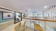 This single-floor space delivers a refined, contemporary work environment that balances open collaboration with private functionality. The layout is anchored by a striking central divider constructed of white perforated paneling topped with a warm wood handrail, creating a visual focal point that organizes the floor without closing it off. Suspended above this feature is a dramatic cluster of slender pendant lights, adding an artistic, sculptural quality to the space.

Light oak flooring runs throughout, lending warmth and continuity across zones. The open lounge areas are furnished with rattan-framed chairs in sage green and cognac leather, arranged to encourage informal gathering. Curated framed artwork in muted lavender and neutral tones lines the white walls, contributing a gallery-like atmosphere.

Glass-partitioned private offices with black steel frames line one side of the floor, offering visual transparency while maintaining acoustic separation. Dedicated desk areas provide additional individual workstations along the perimeter. Built-in open shelving units offer both storage and display space. Large windows allow natural light to filter in, enhancing the overall brightness and airy quality of the interior throughout the day.
