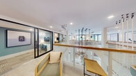This single-floor space delivers a refined, contemporary work environment that balances open collaboration with private functionality. The layout is anchored by a striking central divider constructed of white perforated paneling topped with a warm wood handrail, creating a visual focal point that organizes the floor without closing it off. Suspended above this feature is a dramatic cluster of slender pendant lights, adding an artistic, sculptural quality to the space.

Light oak flooring runs throughout, lending warmth and continuity across zones. The open lounge areas are furnished with rattan-framed chairs in sage green and cognac leather, arranged to encourage informal gathering. Curated framed artwork in muted lavender and neutral tones lines the white walls, contributing a gallery-like atmosphere.

Glass-partitioned private offices with black steel frames line one side of the floor, offering visual transparency while maintaining acoustic separation. Dedicated desk areas provide additional individual workstations along the perimeter. Built-in open shelving units offer both storage and display space. Large windows allow natural light to filter in, enhancing the overall brightness and airy quality of the interior throughout the day.