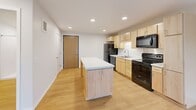 Located at 2800 N Wayzata Blvd in Minneapolis, MN, this single-floor unit offers a clean, modern design with light hardwood flooring carried consistently throughout the space. The open-concept kitchen serves as the heart of the unit, featuring light maple cabinetry, a central island with a white countertop, black appliances including a refrigerator, electric range, microwave, and dishwasher, along with recessed lighting overhead. The kitchen flows seamlessly into the living area, which benefits from large windows that frame views of the surrounding Minneapolis neighborhood and fill the space with natural light. A second room, suitable as a bedroom or flex space, also enjoys generous window views of the tree-lined streetscape outside. This room includes access to a spacious walk-in closet outfitted with built-in shelving and hanging rods, offering ample storage. The bathroom is well-appointed with light maple cabinetry, a single-sink vanity, a walk-in shower with a built-in shelf, and accessibility-friendly grab bars. The overall design aesthetic is clean and contemporary, with neutral white walls, crisp trim, and a bright, airy ambiance throughout.