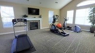 Located at 1802 Kellys Path in Reynoldsburg, Ohio, this single-floor fitness space offers a well-appointed exercise room designed for both functionality and comfort. The room features neutral-toned walls and plush patterned carpeting that provides a cushioned surface underfoot, creating a welcoming environment for workouts. A standout feature is the stone-surround fireplace with a classic white mantel, above which a large wall-mounted flat-screen television is installed — allowing users to stay entertained during exercise sessions. Multiple windows along the walls flood the space with natural light, contributing to an open and energizing atmosphere. The room is equipped with a variety of cardio machines, including a treadmill, elliptical trainers, and a rowing machine, along with a stability ball, accommodating a range of fitness routines. The vaulted ceiling adds a sense of vertical openness, enhancing the spacious feel of the room. The overall ambiance strikes a balance between a practical workout facility and a warm, inviting interior, making it a comfortable space for daily exercise year-round.