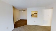 This single-floor space offers a functional and straightforward layout with a seamlessly connected living and dining area. The living area is finished with wall-to-wall carpeting, providing a warm and comfortable feel underfoot, while the adjoining room transitions to dark hardwood-style flooring, adding visual contrast and definition between the two zones. A ceiling fan with a built-in light fixture is centrally positioned overhead, contributing to both air circulation and ambient lighting throughout the space. The walls are painted in a neutral white tone, complemented by a textured popcorn ceiling that adds subtle depth. A notable architectural feature is the pass-through window cut into the dividing wall, which connects the living area directly to the kitchen. This opening offers a glimpse of the kitchen's light wood cabinetry and white refrigerator, allowing for convenient interaction between spaces. The kitchen itself appears compact yet well-organized. The overall ambiance is clean, neutral, and move-in ready, offering a blank canvas that lends itself well to a variety of interior design preferences.