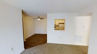 This single-floor space offers a functional and straightforward layout with a seamlessly connected living and dining area. The living area is finished with wall-to-wall carpeting, providing a warm and comfortable feel underfoot, while the adjoining room transitions to dark hardwood-style flooring, adding visual contrast and definition between the two zones. A ceiling fan with a built-in light fixture is centrally positioned overhead, contributing to both air circulation and ambient lighting throughout the space. The walls are painted in a neutral white tone, complemented by a textured popcorn ceiling that adds subtle depth. A notable architectural feature is the pass-through window cut into the dividing wall, which connects the living area directly to the kitchen. This opening offers a glimpse of the kitchen's light wood cabinetry and white refrigerator, allowing for convenient interaction between spaces. The kitchen itself appears compact yet well-organized. The overall ambiance is clean, neutral, and move-in ready, offering a blank canvas that lends itself well to a variety of interior design preferences.