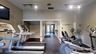 This single-floor fitness center offers a practical and well-organized workout environment suited for a range of exercise routines. The space is outfitted with a selection of cardio equipment, including treadmills and stationary bikes from recognized brands such as Life Fitness, positioned to allow comfortable use and easy movement between machines. The layout is open and functional, with a central corridor leading toward a set of glass doors at the far end, allowing natural light to filter into the space and contributing to an airy, welcoming atmosphere. Durable dark rubber flooring covers the workout area, providing both safety and stability underfoot. Track lighting mounted to the ceiling ensures consistent, bright illumination throughout the facility. A dedicated wall-mounted hook rack is visible along one side, offering convenient storage for personal belongings during workouts. The walls are finished in neutral tones, maintaining a clean and focused environment. A mirrored section enhances the sense of space, while a partially enclosed area along one wall adds structural variety to the layout. Overall, the fitness center presents a straightforward, no-frills approach to exercise, emphasizing accessibility and functionality.