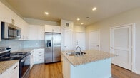 This single-floor space is designed with a clean, contemporary aesthetic throughout. The kitchen serves as a central highlight, showcasing white shaker-style cabinetry that extends to the ceiling, granite countertops with subtle speckled detailing, and a full suite of stainless steel appliances including a gas range, over-the-range microwave, and a side-by-side refrigerator with an in-door ice and water dispenser. A center island provides additional counter space and features a double-basin undermount sink. Light wood-tone flooring runs throughout, lending warmth and visual continuity across all areas. A light blue glass tile backsplash adds a subtle pop of color in the kitchen. The open living area adjacent to the kitchen is bright and airy, benefiting from multiple windows fitted with horizontal blinds that allow for natural light control. A ceiling fan is installed in one of the rooms, enhancing airflow and comfort. The bathroom is finished with a granite-topped white vanity, a large mirror with overhead lighting, and a tub-shower combination clad in large-format marble-style tile. A dedicated laundry closet, accessed through a louvered door, houses a full-size top-load washer and dryer with overhead shelving.