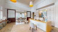 This single-story space is designed with a cohesive coastal aesthetic, blending functional layout with refined interior detailing throughout. The entry area features a prominent wooden reception desk set against a wall of built-in cabinetry, illuminated by warm sconce lighting and accented with curated décor including a coastal landscape artwork and dried pampas grass. Soaring board-and-batten walls and oversized pendant lights with golden interiors reinforce the space's airy, welcoming character.

Adjacent to the reception area, a spacious lounge features polished tile flooring, an array of seating arrangements including woven and upholstered chairs, dual wall-mounted televisions, and decorative wall paneling. Large multi-pane windows flood the area with natural light and offer views of the surrounding exterior.

A set of rich wood-framed sliding barn doors with frosted glass panels opens into a formal dining area centered around a long wooden table with upholstered seating for a large group. A statement industrial pendant light hangs above, while expansive gridded windows frame outdoor views.

A dedicated conference room features a wooden table, wall-mounted display, and direct access to the outdoors. Throughout, polished stone flooring, curated artwork, and layered textiles contribute to a warm, sophisticated ambiance.