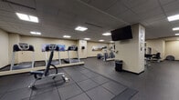 This fitness center at 1717 E Ninth St in Cleveland, Ohio, offers a well-equipped, single-floor exercise facility designed to support a full range of workout routines. The space features a durable rubber tile floor throughout, providing a safe and functional surface for both cardio and strength training activities. The dropped ceiling with recessed panel lighting ensures even, consistent illumination across the entire area.

The cardio section is stocked with multiple treadmills, elliptical trainers, and stair-climbing machines, complemented by wall-mounted flat-screen televisions for entertainment during workouts. Cleveland cityscape photography decorates the walls, adding a local character to the environment. The strength training area includes a selection of Precor selectorized machines, a dumbbell rack with a full range of free weights, medicine balls, and a dedicated flat bench for targeted exercises.

A large wall mirror in the free weights area enhances the sense of space and allows users to monitor their form. The facility also features interior windows and glass partitions that add openness to the layout. Amenities include paper towel dispensers and waste receptacles conveniently placed throughout the space, promoting cleanliness and hygiene. Overall, the gym presents a clean, organized, and functional environment suited for a thorough fitness regimen.