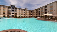 Located at 3200 Peek Rd in Katy, TX, this single-floor residence is part of a modern multi-story community offering exceptional amenities and thoughtfully designed interiors. The community features a expansive resort-style swimming pool surrounded by lounge seating, set against the backdrop of the building's attractive contemporary exterior with varied facade materials and balconies.

Inside, the unit showcases light-toned hardwood-style flooring throughout, complemented by crisp white trim and neutral wall tones that create a clean, modern aesthetic. The entryway features a bold dark front door and recessed lighting, leading into the open-concept living area. A ceiling fan provides comfort in the spacious living and dining area, which benefits from natural light streaming through a large window with outdoor views.

The kitchen is a standout, anchored by a generous granite-topped island with an undermount sink and elegant pendant lighting overhead. Dark espresso cabinetry provides rich contrast, while a full suite of stainless steel appliances — including a refrigerator, electric range, and microwave — ensures modern functionality. A built-in wine rack adds a refined touch. The layout flows seamlessly, making the space ideal for both everyday living and entertaining.
