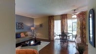 This single-floor residence offers a thoughtfully designed open-concept layout that seamlessly connects the living and dining areas, creating a spacious and cohesive environment. The living room features rich hardwood flooring, a textured ceiling, and a bold accent wall in a cool grey tone that adds depth and character to the space. A plush area rug anchors the seating arrangement, complementing the warm, earthy color palette throughout.

The dining area is positioned adjacent to expansive windows dressed with vibrant, patterned drapes that frame views of the lush greenery outside, allowing abundant natural light to fill the room. A ceiling fan with a light fixture overhead enhances both comfort and illumination. The round dining table with wrought-iron style chairs adds an elegant, classic touch to the space.

Throughout, the design strikes a balance between comfort and style, with decorative wall art, coordinated furnishings, and warm accent lighting contributing to an inviting ambiance. The large sliding glass door or window panel further blurs the boundary between indoors and outdoors, making the space feel bright, airy, and connected to its natural surroundings.