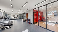 Located at 250 Plaza Circle in Mundelein, IL, this well-appointed fitness center spans a single floor and offers a comprehensive workout environment thoughtfully divided into two dedicated exercise rooms. The main exercise area features durable speckled rubber flooring and is bathed in natural light from expansive floor-to-ceiling windows that offer outdoor views of the surrounding community. This space is equipped with a row of treadmills and elliptical machines, a full dumbbell rack, and a dedicated strength training zone outfitted with power racks, barbells, weight plates, and a cable machine. Floor-to-ceiling mirrors line the strength area walls, amplifying the sense of space and allowing users to monitor their form. Sleek linear LED lighting runs along the ceiling throughout, providing consistent, bright illumination. A separate enclosed studio, distinguished by its bold red accent wall and glass partition walls, houses spin bikes and a wall-mounted television, making it ideal for cycling classes or focused cardio sessions. Light hardwood flooring and a full mirrored wall complete the studio's clean, energetic aesthetic. The overall design is modern and functional, with a neutral white-and-gray palette accented by striking red details that lend the space a dynamic, motivating atmosphere.
