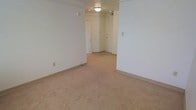 This single-floor space presents a clean, minimalist layout with a neutral color palette throughout. The interior is finished with wall-to-wall beige carpeting that extends across the main living area, providing a warm and cohesive feel underfoot. The walls are painted in a crisp off-white tone, contributing to a bright and open ambiance despite the absence of windows visible in the primary view. Overhead, a flush-mount ceiling light fixture provides functional illumination, supplemented by additional recessed lighting in the connecting hallway area. The layout features multiple doorways leading to adjacent rooms or entry points, suggesting a well-connected floor plan with clear transitions between spaces. Exposed conduit or track elements along the upper wall add a subtle utilitarian character to the space. Baseboards run cleanly along the perimeter, and electrical outlets are positioned conveniently along the walls. The overall design is straightforward and practical, offering a neutral canvas well-suited for a variety of uses. The space feels open and uncluttered, with the hallway corridor lending depth and a sense of flow to the overall layout.