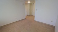 This single-floor space presents a clean, minimalist layout with a neutral color palette throughout. The interior is finished with wall-to-wall beige carpeting that extends across the main living area, providing a warm and cohesive feel underfoot. The walls are painted in a crisp off-white tone, contributing to a bright and open ambiance despite the absence of windows visible in the primary view. Overhead, a flush-mount ceiling light fixture provides functional illumination, supplemented by additional recessed lighting in the connecting hallway area. The layout features multiple doorways leading to adjacent rooms or entry points, suggesting a well-connected floor plan with clear transitions between spaces. Exposed conduit or track elements along the upper wall add a subtle utilitarian character to the space. Baseboards run cleanly along the perimeter, and electrical outlets are positioned conveniently along the walls. The overall design is straightforward and practical, offering a neutral canvas well-suited for a variety of uses. The space feels open and uncluttered, with the hallway corridor lending depth and a sense of flow to the overall layout.