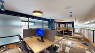 Located at 1010 E Orange St in Tempe, Arizona, this modern office suite offers a thoughtfully designed, single-floor workspace tailored for productivity and collaboration. The open floor plan accommodates multiple workstation clusters equipped with privacy-screened desks and monitors, providing focused individual work areas within a shared environment.

Bold navy blue accent walls contrast sharply with white ceilings and clean architectural lines, creating a professional yet energetic atmosphere. Underfoot, a striking geometric-patterned carpet in orange, navy, white, and gray ties the space together with a contemporary flair. Overhead, a combination of round flush-mount drum pendants and vibrant teal cylindrical pendant lights delivers warm, layered illumination throughout.

Semi-private meeting areas are enclosed by floor-to-ceiling frosted and clear glass partition walls with dark framing, offering visual separation while maintaining an open feel. A built-in kitchen counter with bar-height seating and cabinetry provides a casual collaboration zone. Large windows along the perimeter flood the space with natural light and offer views of the surrounding building.

A perforated metal-railed mezzanine walkway overlooks the lower building atrium, adding depth and architectural interest. The overall design balances open collaboration with private focus zones, making this a highly functional and visually dynamic workspace.