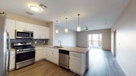 This single-floor unit showcases a clean, contemporary design throughout. The open-concept layout seamlessly connects the kitchen and living area, creating a bright, airy atmosphere enhanced by wood-look flooring and ceiling fans. The kitchen is well-appointed with white shaker cabinetry, quartz countertops, stainless steel appliances including a gas range, microwave, dishwasher, and refrigerator, complemented by a striking herringbone-patterned tile backsplash. A peninsula with pendant lighting above provides additional prep space and casual seating, while sliding doors at the far end of the living area lead to a private balcony with open views.

The bedroom is generously proportioned, featuring plush carpeting, a ceiling fan, and a window that draws in natural light and exterior views. The bathroom offers a dual-sink vanity with white cabinetry, quartz countertops, and framed mirrors with wall-mounted lighting, alongside a glass-enclosed walk-in shower finished in neutral tile. A dedicated laundry area houses a stacked washer and dryer unit, adding everyday convenience. The overall palette of warm neutrals, modern fixtures, and consistent finishes creates a cohesive and functional living environment.