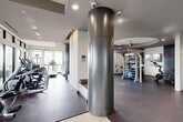 Located at 2800 Kirby Drive in Houston's Upper Kirby neighborhood, this fitness center offers a generously sized, single-floor exercise space designed with both functionality and aesthetics in mind. The layout is thoughtfully divided into distinct workout zones, allowing for a variety of training modalities. Floor-to-ceiling windows and large glass walls flood the interior with natural light while offering views of the surrounding cityscape, creating an energizing atmosphere for workouts. The cardio zone is well-appointed with a row of Matrix treadmills positioned along the windowed walls, complemented by ellipticals and stationary bikes on durable rubber flooring. A dedicated functional training area features a multi-use training tower with TRX suspension straps, resistance bands, and storage for accessories such as medicine balls and exercise mats. A distinctive curved ceiling detail with a ceiling fan adds architectural interest and visual depth to the open training floor. The space is finished with polished concrete and dark rubber flooring, recessed lighting, and a wall-mounted flat-screen television. Large sliding glass doors connect seamlessly to an adjacent lounge area, enhancing the sense of openness throughout this well-designed fitness facility.
