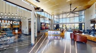 This single-story amenity center makes a bold architectural statement with its aviation-inspired design theme carried throughout. The expansive main lobby commands attention with soaring ceilings, dramatic floor-to-ceiling glass curtain walls, and polished concrete floors inlaid with linear LED light strips that radiate outward from the entrance. Oversized industrial-style ceiling fans and warm metallic ceiling finishes lend a sophisticated, industrial-modern aesthetic. A centrally positioned illuminated reception island in warm wood tones serves as the focal point of the space, flanked by a lounge area with plush sectional sofas, leather club chairs, and a large multi-screen video wall featuring aviation-themed artwork. Natural light floods the interior, offering views of the surrounding palm-lined outdoor spaces. A dedicated dining area showcases striking aviation mural wallcoverings, glass-top tables, and direct access to the outdoor patio. The fitness center is thoughtfully divided into two zones: a cardio and functional training studio with cycling bikes, TRX suspension systems, medicine balls, and kettlebells alongside views of the outdoors; and a larger strength training area with cable machines, free weights, a heavy punching bag, and cardio equipment. Full-length mirrors and rubber flooring complete the well-appointed gym.