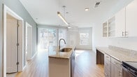This single-floor residence showcases a contemporary open-concept design that seamlessly connects the kitchen and living areas. The kitchen is a functional centerpiece, equipped with a granite-topped island featuring an undermount sink, dark cabinetry paired with white upper cabinets, open shelving, and black appliances including a range, microwave, and refrigerator. Light wood-look flooring flows throughout the main living spaces, contributing to a clean, modern aesthetic.

The living area is notably bright, benefiting from multiple windows and a full-height sliding glass door fitted with vertical blinds that open to an outdoor area. A ceiling fan with a light fixture provides additional comfort and airflow. The open layout creates a generous sense of space, while accent walls in a soft sage green add subtle visual interest.

The bedroom is carpeted and filled with natural light from a large window, offering a quiet, comfortable retreat. The bathroom is finished with large-format tile flooring and matching wall tile in the walk-in shower, complemented by a granite vanity, white cabinetry, and backlit mirrors. Overall, the unit presents a bright, well-appointed interior with consistent modern finishes throughout.
