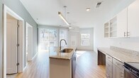 This single-floor residence showcases a contemporary open-concept design that seamlessly connects the kitchen and living areas. The kitchen is a functional centerpiece, equipped with a granite-topped island featuring an undermount sink, dark cabinetry paired with white upper cabinets, open shelving, and black appliances including a range, microwave, and refrigerator. Light wood-look flooring flows throughout the main living spaces, contributing to a clean, modern aesthetic.

The living area is notably bright, benefiting from multiple windows and a full-height sliding glass door fitted with vertical blinds that open to an outdoor area. A ceiling fan with a light fixture provides additional comfort and airflow. The open layout creates a generous sense of space, while accent walls in a soft sage green add subtle visual interest.

The bedroom is carpeted and filled with natural light from a large window, offering a quiet, comfortable retreat. The bathroom is finished with large-format tile flooring and matching wall tile in the walk-in shower, complemented by a granite vanity, white cabinetry, and backlit mirrors. Overall, the unit presents a bright, well-appointed interior with consistent modern finishes throughout.