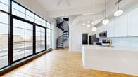 This two-story penthouse at 268 Alexander St in Rochester, NY offers a striking blend of industrial character and modern finishes across two thoughtfully designed floors. The main level features an open-plan living, dining, and kitchen area anchored by a bold black spiral staircase that serves as a dramatic architectural centerpiece. Soaring ceilings with exposed ductwork and structural beams lend an industrial loft aesthetic, while large floor-to-ceiling factory-style windows flood the space with natural light and frame rooftop views of the surrounding cityscape. A set of French-style doors opens directly onto a private outdoor terrace, extending the living space outdoors. The kitchen is well-appointed with white shaker cabinetry, subway tile backsplash, stainless steel appliances, quartz countertops, and a central island. Warm natural wood flooring flows throughout both levels, complementing the bright white walls. The spacious primary bedroom features a bank of windows with rooftop views, while the bathrooms are finished in a clean, contemporary style with white vanities and neutral tile. A second bedroom offers access to its own outdoor space. Three full bathrooms ensure convenience across the penthouse.