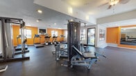 Located at 141 S Jamaica Way in Aurora, Colorado, this fitness center offers a well-equipped, single-floor workout facility designed to support a full range of exercise routines. The space is defined by its bold orange accent walls, textured ceilings, and dark rubber flooring, creating an energetic and functional atmosphere throughout.

The cardio area is outfitted with multiple treadmills, elliptical trainers, and a stationary cycling bike, all positioned along windowed walls that allow generous natural light to fill the room. A wall-mounted television provides entertainment during cardio sessions. The strength training section features a multi-station cable machine, a Smith machine with a rack, a dedicated bench press setup, and a collection of free weights and dumbbells, all reflected in large wall mirrors that enhance the sense of space.

An open stretch and functional training area provides ample floor space for bodyweight exercises and mobility work. The facility benefits from multiple windows along several walls, offering views of the surrounding outdoor areas and maintaining a bright, airy environment. Recessed lighting and wall-mounted sconces supplement natural light throughout the day. A ceiling fan adds comfort to portions of the space, while direct exterior access connects the fitness center to outdoor amenities, including what appears to be a pool area, making this a comprehensive and convenient fitness destination.