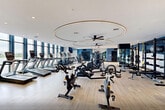 Located on Goldsborough Drive in Bayonne, NJ, this fitness center offers a generously sized, single-floor exercise room designed with both function and aesthetics in mind. The space features light hardwood-style flooring throughout, crisp white ceilings with recessed lighting, and bold navy blue accent walls that lend a modern, energetic atmosphere.

Large floor-to-ceiling windows line the perimeter, flooding the space with natural light and providing scenic outdoor views, including waterfront scenery. Ceiling fans with circular decorative lighting fixtures enhance airflow and visual appeal, while full-length mirrors along multiple walls amplify the sense of openness and assist users during workouts.

The cardio zone is extensively equipped with treadmills, ellipticals, stair climbers, stationary bikes, and Matrix rowing machines. The strength training area includes a power rack with TRX suspension systems, cable machines, a full dumbbell rack, adjustable benches, and additional selectorized equipment. A dedicated functional training column and a wall-mounted TV add further utility to the space.

Additional amenities include a built-in water fountain station, ample open floor space for stretching or functional movement, and private room access visible through glass-paneled doors. The overall design balances professional-grade equipment with an inviting, well-lit environment suited for a comprehensive fitness experience.