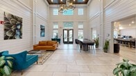 Located at 150 Country Squire Dr in Cromwell, Connecticut, this single-floor community clubhouse offers a generously sized, thoughtfully designed gathering space that seamlessly blends elegance with everyday functionality. The entrance foyer makes an immediate impression with soaring double-height ceilings clad in white shiplap paneling, large transom windows that flood the space with natural light, and a striking geometric chandelier that anchors the room. Large-format stone tile flooring extends throughout the entrance area, lending a clean, sophisticated foundation. A dramatic floor-to-ceiling stacked stone fireplace serves as the room's focal point, topped with a wall-mounted television and flanked by plush blue accent chairs, creating an inviting lounge setting. Transitioning deeper into the space, wood-look plank flooring introduces a warmer, more casual aesthetic. This area is outfitted with a high-top bar counter with seating, casual seating vignettes, and access to a coffee station. A dedicated co-working zone features built-in desk nooks with navy blue paneling and individual task lighting. The game room area includes a shuffleboard table, curated wall art, and flexible seating arrangements. Throughout, recessed lighting, contemporary furnishings, and carefully selected artwork contribute to a polished yet welcoming atmosphere.