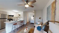 This well-designed single-floor unit offers a cohesive open-concept layout that seamlessly connects the kitchen, dining, and living areas. The kitchen is outfitted with white cabinetry, stainless steel appliances including a refrigerator, microwave, and electric range, and a light quartz-topped peninsula with bar seating — ideal for casual dining or entertaining. Wood-look flooring runs throughout, lending warmth and continuity across the space. The living area is bright and airy, benefiting from a large window that invites natural light, and is anchored by a ceiling fan overhead. A louvered bi-fold divider separates the bedroom from the main living space, offering both privacy and an open feel when desired. The bedroom is comfortably sized and also features a ceiling fan. A dedicated workspace with a desk and chair is neatly integrated near the kitchen, making the unit well-suited for remote work. The bathroom includes a full shower-tub combination with clean white tile, and a separate vanity area with a framed mirror and white basin sink. Throughout the unit, neutral tones and consistent finishes create a clean, functional, and inviting atmosphere.