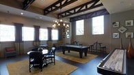 This single-floor recreational lounge is designed to offer a sophisticated yet relaxed environment for social gatherings and leisure activities. The space is anchored by a prominent pool table at its center, surrounded by an elegant geometric-patterned area rug in warm gold and neutral tones that defines the gaming zone. A round dining or game table with cushioned chairs occupies one side of the room, providing an intimate setting for card games or casual conversation.

Architecturally, the space features striking exposed dark wood truss detailing along the ceiling, adding a rustic yet refined character. A wrought-iron chandelier with candle-style lighting hangs centrally, enhancing the warm ambiance. High clerestory windows and full-length draped windows flood the room with abundant natural light while offering views of the surrounding exterior. Tall bar-height tables with stools are positioned near the windows, creating additional casual seating options.

The walls are finished in a soft neutral tone, complemented by framed artwork. Dark hardwood-style flooring grounds the space, contrasting beautifully with the warm rug beneath. Overall, the lounge balances functionality with an inviting, upscale aesthetic.