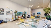 This thoughtfully designed single-floor amenity lounge offers a generously proportioned open-plan layout that seamlessly integrates multiple functional zones. The space is defined by striking floor-to-ceiling curtain wall glazing along one full facade, framing panoramic urban views and flooding the interior with abundant natural light.

The dining and collaboration zone features a large rectangular wooden table surrounded by a mix of upholstered chairs in rich blue and warm orange tones. A wall-mounted display screen and a low credenza with open shelving—stocked with a coffee station and accessories—serve this area efficiently. Geometric pendant lighting adds a modern architectural accent overhead.

Transitioning deeper into the space, a relaxed lounge area is anchored by a bold, abstract area rug in muted greens, yellows, and neutrals. Accent seating in mustard yellow and teal velvet is arranged around the rug, complemented by high-back privacy chairs and small bistro tables. A full-length shuffleboard table provides a playful recreational element.

The walls serve as a curated gallery, displaying vinyl record artwork, framed photographs, decorative guitars, and sculptural wall pieces. Open black metal shelving units house lush indoor plants, books, and decorative objects, adding warmth and texture throughout. The overall ambiance is energetic yet welcoming, balancing functionality with creative, eclectic design.