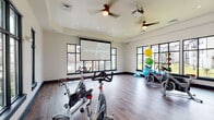 This dedicated fitness studio is designed to support a wide range of workout routines in a bright, open environment. The space features rich hardwood-style flooring throughout, crisp white walls with dark-trimmed accents, and recessed lighting complemented by ceiling fans that keep the room comfortable during high-intensity sessions. Large, black-framed windows line multiple walls, flooding the interior with natural light and offering views of the surrounding grounds and pool area, creating an energizing atmosphere for exercise. A large retractable projection screen serves as a central focal point, enabling instructor-led virtual classes and guided workout sessions directly within the studio. Multiple stationary spin bikes are arranged across the open floor, accommodating group cycling classes with ease. A well-stocked equipment corner houses an assortment of stability balls in various sizes, a BOSU balance trainer, resistance bands, yoga mats, and medicine balls, supporting diverse functional fitness and flexibility training. A wall-mounted display panel provides additional interactive capabilities, while posted informational signage keeps users informed of studio guidelines. The spacious, open layout offers ample room for movement-based workouts beyond cycling, making this studio a versatile and fully equipped fitness destination.
