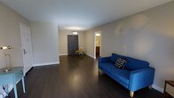 This single-floor space presents a clean, modern aesthetic defined by its rich, dark hardwood flooring that extends throughout the open living area, creating a striking contrast against the neutral gray-toned walls and crisp white baseboards. The living room is generously proportioned, offering ample space for flexible furniture arrangements. A bold blue sofa anchors one side of the room, adding a vibrant pop of color to the otherwise understated palette. Overhead, a simple pendant light fixture provides central illumination, complementing the room's understated modern style. Toward the back of the space, a decorative panel and a small accent chair add a touch of personality without overcrowding the open floor plan. A doorway leads to what appears to be a bathroom, suggesting a practical and well-organized layout. The overall ambiance is bright, airy, and move-in ready, with a minimalist sensibility that allows for easy personalization. The combination of dark flooring and light walls gives the space a balanced, welcoming character well-suited to a variety of living styles.