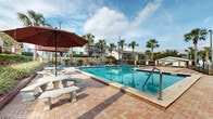 Located at 840 S Grand Hwy in Clermont, Florida, this outdoor amenity area centers around a generously sized swimming pool with clear, blue-tinted water and tiled trim detailing. The pool is framed by an expansive brick paver deck that provides ample space for relaxation and recreation. Surrounding the pool, a collection of blue lounge chairs and shaded umbrella tables offer comfortable spots for sunbathing and socializing under the Florida sky.

Tall palm trees are strategically planted throughout the area, lending a tropical ambiance and natural shade. Lush, manicured landscaping with ornamental plants and mulched garden beds lines the perimeter, complemented by decorative brick retaining walls and wrought iron fencing that define the space with a refined, secure enclosure.

A broad, multi-tiered brick staircase serves as a grand entrance to the pool area, adding an architectural focal point to the grounds. An adjacent single-story structure with a shingle roof houses convenience amenities, including vending machines, providing easy access for guests. Decorative column pillars with lantern-style lighting mark the gated entry, adding a welcoming touch. The surrounding multi-story residential buildings with balconies overlook the pool area, completing this well-appointed Florida outdoor retreat.