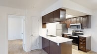 This single-floor residence offers a thoughtfully designed layout encompassing a welcoming entrance, an open-concept living and kitchen area, one generously sized bedroom, and a dedicated laundry room. The living room is bright and open, featuring plush wall-to-wall carpeting, neutral white walls, and a sliding glass door that fills the space with natural light and provides direct access to a private outdoor area. The kitchen is a functional centerpiece of the home, featuring rich dark-espresso cabinetry, a peninsula with a white countertop and built-in sink, and a full suite of stainless steel appliances including a refrigerator, electric range, and microwave. A skylight overhead bathes the kitchen in additional natural light, while tile flooring adds a clean, durable finish. The spacious primary bedroom is carpeted and bright, with a window offering natural light and direct access to a full bathroom. The laundry room includes a stacked washer and dryer unit with wire shelving for added storage convenience. Throughout the residence, the clean white walls, consistent flooring choices, and practical layout create a cohesive and comfortable living environment.