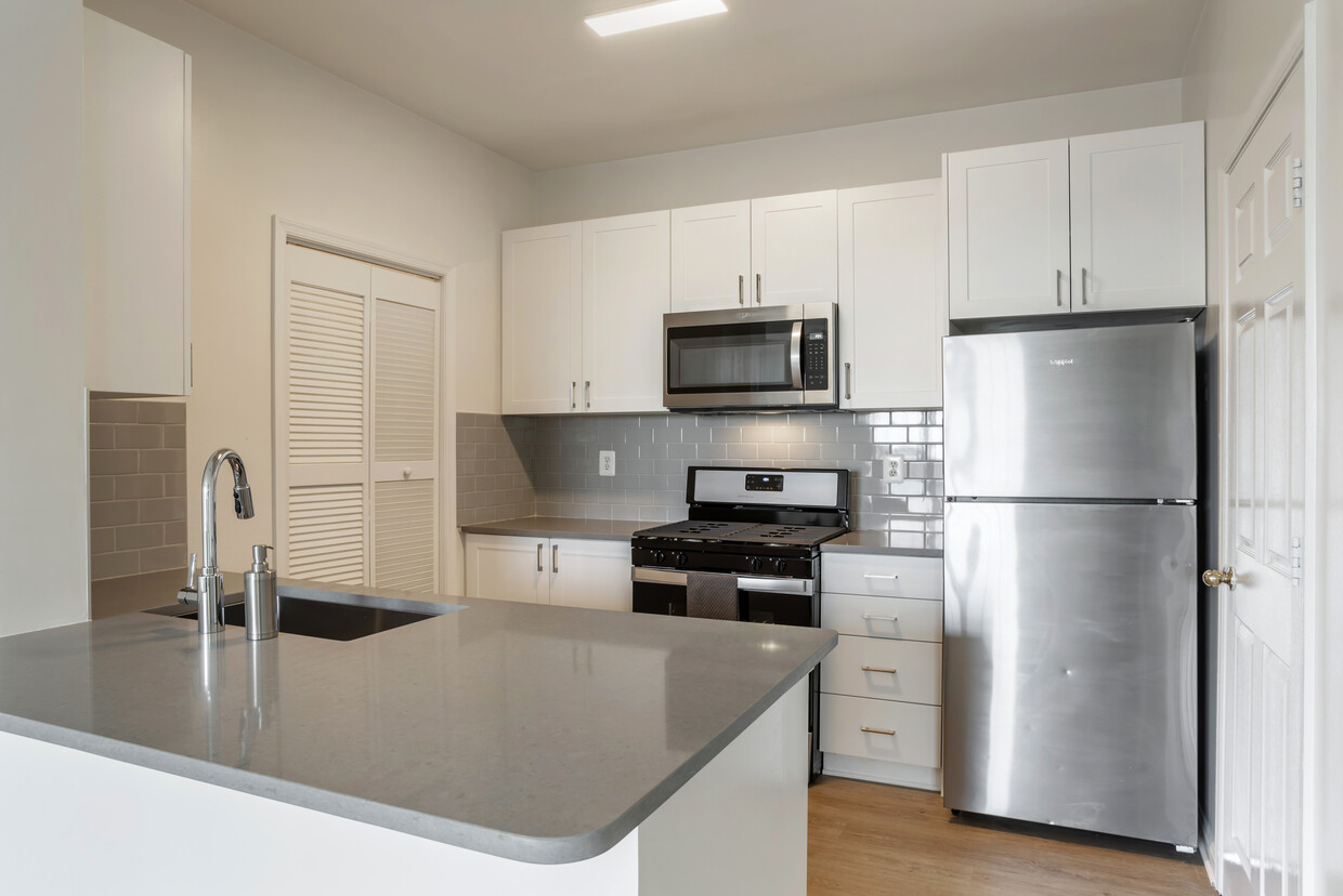 Cocina renovada del Paquete II con gabinetes blancos, encimeras de cuarzo gris, salpicadero de azulejos, electrodomésticos de acero inoxidable y pisos de superficie dura - Avalon Reston Landing