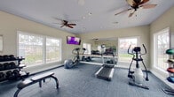This dedicated fitness center is designed to support a comprehensive workout routine in a bright and functional environment. The space is finished with durable, dark blue carpet tile flooring throughout, providing a practical and comfortable surface for exercise. Multiple large windows line the walls, flooding the room with natural light and offering views of the surrounding outdoor areas, including a glimpse of a pool and landscaped grounds. Ceiling fans are positioned throughout to ensure consistent airflow and comfort during workouts. The cardio area is equipped with treadmills, elliptical machines, and a rowing machine, offering a variety of aerobic exercise options. A full-wall mirror with a mounted wooden ballet bar runs along one side of the room, enhancing the sense of space and providing a useful reference point for form and technique. A wall-mounted flat-screen television is positioned for easy viewing from the cardio equipment. A dedicated free weights section features a multi-tier dumbbell rack stocked with a range of weights, accompanied by a weight bench for strength training. An illuminated exit sign and a clearly marked door provide convenient access, reinforcing the safety and functionality of the space.