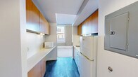 Located at 218 6th Ave in the heart of downtown Des Moines, Iowa, this efficient single-floor unit offers a well-considered layout that makes smart use of every square foot. The galley-style kitchen is a standout feature, lined on both sides with warm honey-toned wood cabinetry that extends to the ceiling, providing ample storage. White appliances — including a refrigerator, electric range, and microwave — complement the clean white countertops, while dark tile flooring grounds the space with a bold contrast. A central architectural column with a dark navy accent visually anchors the transition between the kitchen and the open living area, which features neutral wall-to-wall carpeting that adds warmth underfoot. Large double-hung windows in the main living area flood the space with natural light and frame views of the surrounding downtown skyline, enhancing the sense of openness. The bathroom is clean and functional, featuring a tub-shower combination with crisp white surround panels and chrome fixtures. The natural wood entry door adds an inviting, warm touch to the overall modern and utilitarian aesthetic of this downtown unit.