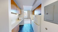 Located at 218 6th Ave in the heart of downtown Des Moines, Iowa, this efficient single-floor unit offers a well-considered layout that makes smart use of every square foot. The galley-style kitchen is a standout feature, lined on both sides with warm honey-toned wood cabinetry that extends to the ceiling, providing ample storage. White appliances — including a refrigerator, electric range, and microwave — complement the clean white countertops, while dark tile flooring grounds the space with a bold contrast. A central architectural column with a dark navy accent visually anchors the transition between the kitchen and the open living area, which features neutral wall-to-wall carpeting that adds warmth underfoot. Large double-hung windows in the main living area flood the space with natural light and frame views of the surrounding downtown skyline, enhancing the sense of openness. The bathroom is clean and functional, featuring a tub-shower combination with crisp white surround panels and chrome fixtures. The natural wood entry door adds an inviting, warm touch to the overall modern and utilitarian aesthetic of this downtown unit.