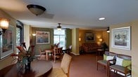 This single-floor space offers an open, flowing layout that seamlessly connects a dining area and a living room, creating a cohesive and functional environment. The interior is finished in warm earth tones, with olive green and tan walls that lend a cozy, welcoming ambiance throughout.

The dining area features a rounded wood table surrounded by cushioned wooden chairs, positioned near a window with horizontal blinds that allows natural light to filter into the space. A ceiling fan with a light fixture is mounted above, contributing to both comfort and airflow. Framed landscape artwork adorns the walls, adding a decorative touch.

The adjacent living room is carpeted in neutral tones and furnished with a patterned upholstered sofa and accompanying accent chairs. Additional ceiling fans extend across this area as well. Wall sconces and table lamps provide warm ambient lighting, complementing the natural light. A side table with decorative greenery adds a natural element to the room.

Overall, the space presents a practical and comfortable single-level layout with consistent décor, warm lighting, and a relaxed residential atmosphere.