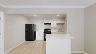 This efficiently designed single-floor unit offers a practical and well-organized layout comprising a living room, kitchen, bedroom, bathroom, and closet. The entryway welcomes you with light tile flooring and crown molding accents, setting a clean, neutral tone throughout. The kitchen is a functional highlight, featuring white shaker-style cabinetry with brushed nickel hardware, a marble-style countertop, a double stainless steel sink, a black refrigerator with water dispenser, a black range, and a peninsula that provides additional prep and counter space. Tile flooring extends through the kitchen and living areas for easy maintenance. The bedroom offers plush carpeting, a ceiling fan with light fixture, a window with blinds allowing natural light, and direct access to a dedicated closet outfitted with dual shelving rods and carpet flooring — ideal for organized storage. The full bathroom features tile flooring, a white vanity with marble-style countertop, and a tub-shower combination with white tile surround. A window unit provides climate control in one of the rooms, and the overall neutral color palette throughout creates a bright, clean ambiance suited for comfortable everyday living.