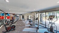This spacious fitness center is thoughtfully designed to support a full range of workout routines. The main exercise area features patterned carpet flooring in neutral tones of gray and blue, recessed lighting, and notably high ceilings that create an open, energizing atmosphere. Large floor-to-ceiling glass panels along one wall allow abundant natural light to filter in while offering views of the outdoors.

The facility is comprehensively equipped with cable machines, weight benches, a rack of dumbbells, medicine balls, resistance bands, stability balls, and cardio equipment including stationary bikes. Full-length mirrors line sections of the wall, enhancing the sense of space and allowing users to monitor their form. A large-format athletic photo mural adds a dynamic, motivational focal point to the space, while a wall-mounted flat-screen television provides additional entertainment.

A dedicated area with open floor space accommodates functional training and stretching. The facility also includes two restrooms, each distinctively designed. One features white and gold hexagonal marble-style tile with a backlit mirror and marble flooring, while the other showcases a dramatic three-dimensional metallic geometric tile wall, a floating vanity, and a backlit mirror, both reflecting a high standard of finish throughout.