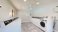 Located at 2700 Iowa Ave in Riverside, California, this shared laundry facility is a efficiently designed, single-floor space offering a comprehensive suite of laundry amenities. The room is outfitted with multiple top-loading washers and front-loading dryers arranged neatly along the walls, providing ample capacity for residents. Light wood-look laminate flooring runs throughout, lending a clean and modern aesthetic to the space. The interior is finished with neutral light gray walls and white trim, creating a bright, tidy atmosphere. Recessed lighting and a centrally mounted ceiling fan ensure the room remains well-lit and comfortably ventilated. A built-in countertop along one wall offers convenient folding and sorting space, complemented by white cabinetry below for additional utility. Multiple windows allow natural light to enter and provide views to the exterior, further enhancing the open feel of the room. Informational signage and laundry usage guides are posted on the walls for user reference. A wall-mounted air conditioning unit provides additional climate control. The space is thoughtfully organized and well-maintained, making it a practical and comfortable shared amenity for residents of the property.