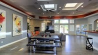 This single-floor space showcases a contemporary open-plan design that seamlessly blends style and functionality. The interior is immediately distinguished by its striking tray ceiling, which is fitted with multiple skylights that flood the room with abundant natural light, complemented by recessed lighting and a modern ceiling fan. Bold red accent bands along the upper walls add a vivid contrast to the otherwise neutral gray palette, creating a dynamic and energetic atmosphere. A standout stacked-stone fireplace anchors one side of the room, providing a warm, textural focal point that pairs beautifully with a large flat-screen television mounted above it. The living and dining areas flow naturally into one another, furnished with a substantial dining table paired with sleek gray upholstered chairs and a live-edge bench, alongside a lounge area featuring bold orange accent seating. Polished concrete floors run throughout, lending an industrial yet refined character to the space. Floor-to-ceiling windows and glass doors along the far wall bathe the interior in additional daylight while offering views of lush greenery outside, creating a seamless indoor-outdoor connection. The overall ambiance is modern, welcoming, and well-appointed — suited for both social gatherings and relaxed everyday use.